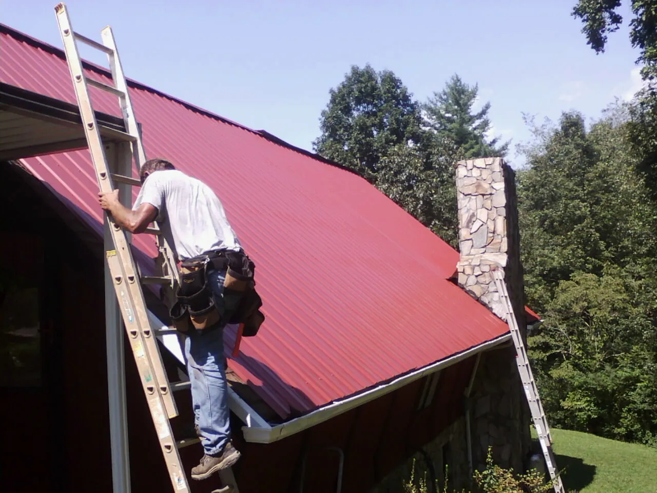 Roofer on a ladder beside a red metal roof and stone chimney — Soffit & Fascia Repair by Roofing Brothers in Hanover